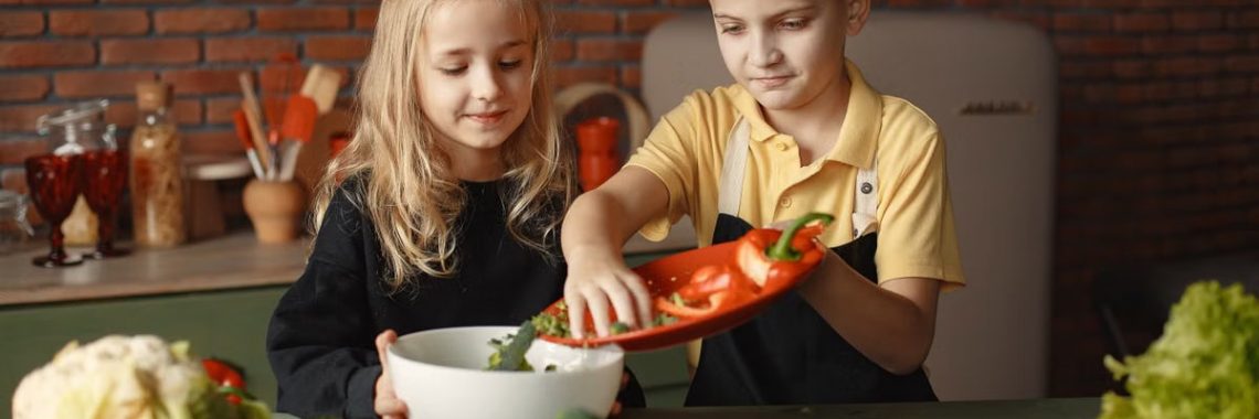 Little girl and boy mixing cutted pepper with other fresh ingredients for salad in big bowl while cooking at table with various fresh vegetables and fruits
